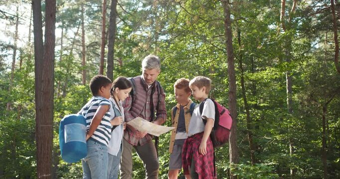 Caucasian man teacher standing in forest and examining route plan with her pupils. Male couch resting in wood with kids while having walk outdoor and looking at map. Tourism concept