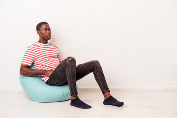 Fototapeta premium Young African American man sitting on a puff isolated on white background confused, feels doubtful and unsure.