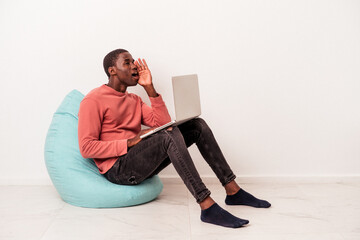 Young African American man sitting on a puff using laptop isolated on white background shouting and holding palm near opened mouth.