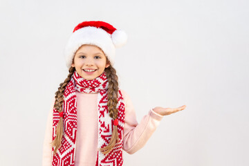 A little girl in a Christmas hat and scarf points her palm to the side and smiles.