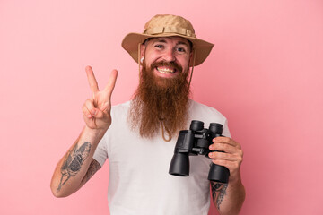 Young caucasian ginger man with long beard holding binoculars isolated on pink background showing number two with fingers.