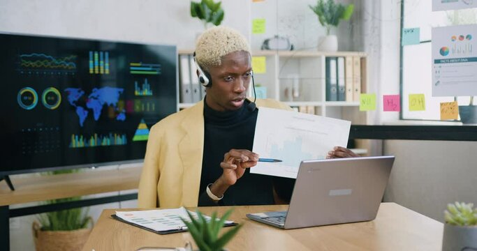 Likable confident responsible skilled african american male office worker in headset explaining histogram for online colleagues through laptop during video chat