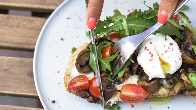 Woman hands cut canape with pouched egg mushrooms tomatoes and ruccola on piece of bread with knife and fork extreme closeup