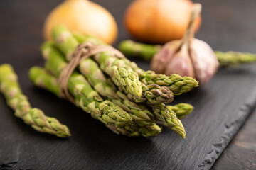 Bunch of fresh green asparagus, garlic, onion on slate board on black concrete background. Side view, close up, selective focus.