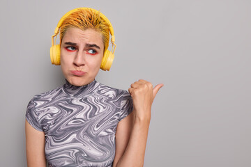 Upset teenage girl with dyed yellow hair purses lips and points thumb away at blank copy space beinng dissatisfied with something wears casual t shirt listens music in wireless stereo headphones