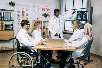 Obraz premium Group of multiracial medical specialists sitting together at office desk and listening african colleague that pointing on x ray scan. Competent doctors examining results of patient analysis.