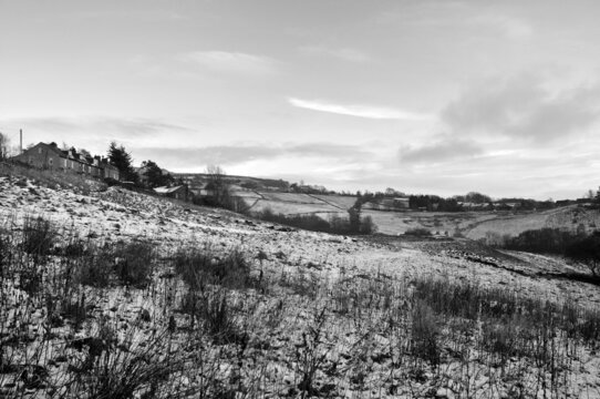 monochrome view of a snow covered narrow country lane with a row of stone houses surrounded by fields at hurst lane in hebden bridge surrounded by west yorkshire pennine landscape