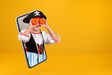 A child in a carnival costume of a pirate looks from a smartphone. Little girl on Halloween online on a mobile phone screen and holding pumpkins instead of eyes. Copy space.