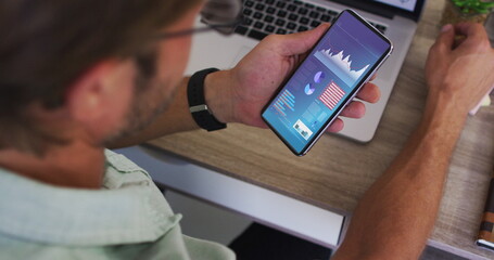 Caucasian man sitting at desk using smartphone with statistics on screen