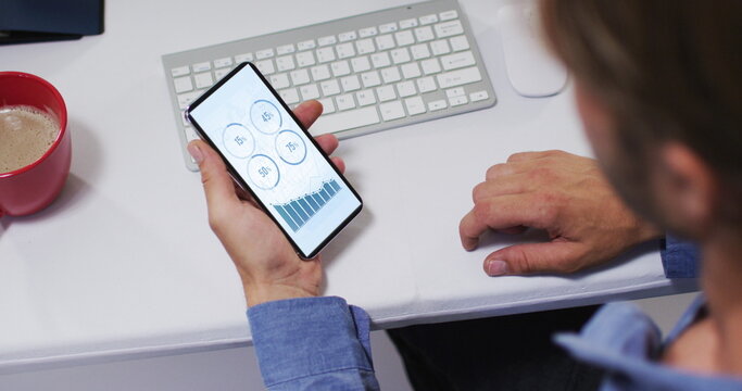 Caucasian man sitting at desk using smartphone with statistics on screen
