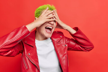 Indoor shot of extraordinary hipster girl laughs happily tries to hide her face has green hairstyle...