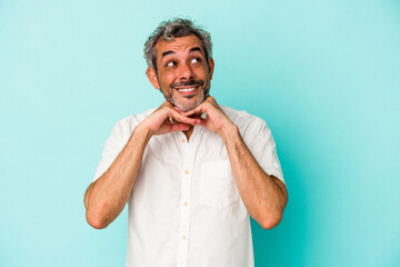 Middle age caucasian man isolated on blue background  keeps hands under chin, is looking happily aside.