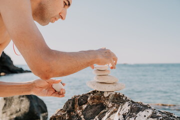 Man bilds stones pyramid on the seashore on a sunny day on the blue sea background. Happy holidays. Pebble beach, calm sea, travel destination. Concept of happy vacation on the sea, meditation, spa