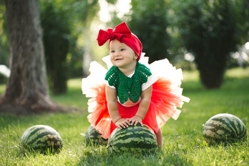 Small girl is sitting on the green grass in a red tulle skirt with watermelons.