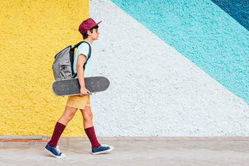 Boy with backpack and skateboard walking by multi colored wall