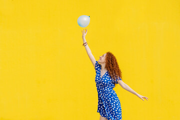 Carefree redhead woman playing with balloon in front of yellow wall