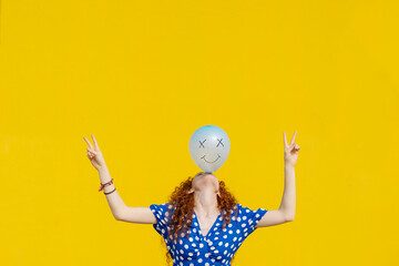 Carefree woman balancing balloon with anthropomorphic smiley face in front of yellow wall
