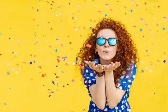 Beautiful woman blowing confetti in front of yellow wall
