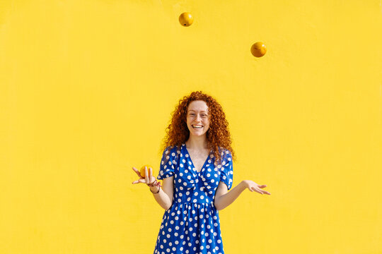 Carefree woman juggling orange fruits in front of yellow wall