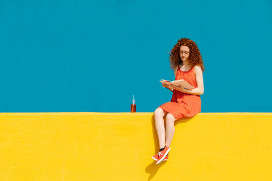 Young redhead woman reading book while sitting on yellow retaining wall