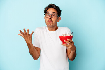 Young mixed race man holding cereals isolated on blue background shrugs shoulders and open eyes...