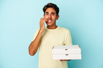 Young mixed race man holding pizzas isolated on blue background biting fingernails, nervous and...