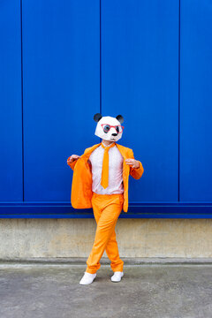 Man wearing vibrant orange suit and panda mask standing confidently against blue wall