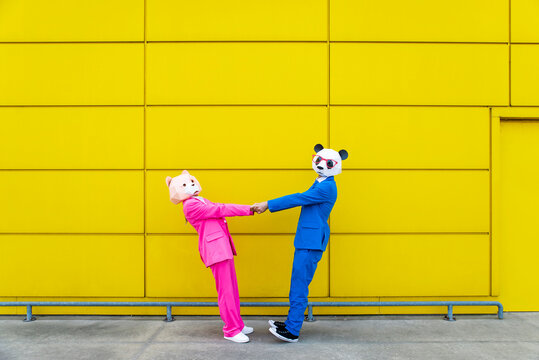 Man and woman wearing vibrant suits and bear masks holding hands against yellow wall