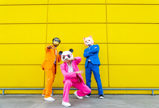 Three adults wearing vibrant suits and animal masks posing together in front of yellow wall