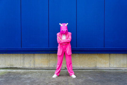 Woman wearing vibrant pink suit and hippo mask standing in front of blue wall with crossed arms