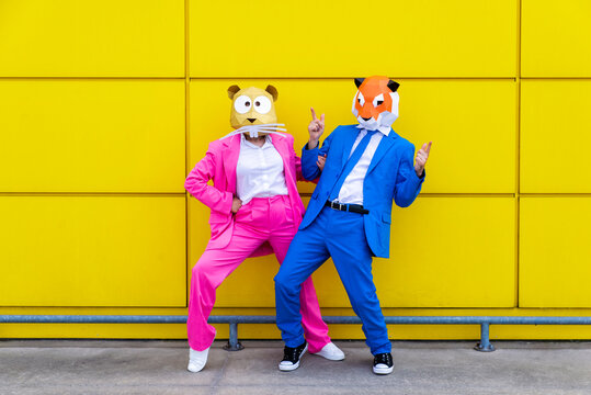Man and woman wearing vibrant suits and animal masks posing together against yellow wall