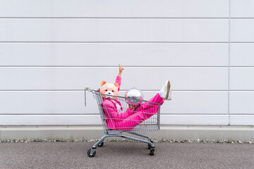 Woman wearing vibrant pink suit and bear mask sitting in shopping cart with disco ball in hands