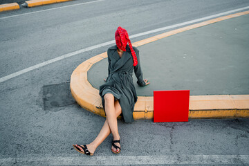 Woman covering face with red stocking while sitting by placard on road