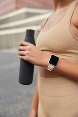 Cropped shot of unrecognizable fit woman dressed in tracksuit has break after intensive workout holds bottle of fresh water uses wearable digital smartwatch for results tracking poses outside