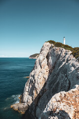 Fototapeta na wymiar Ionian sea island steep rocky cliffs with lighthouse on top in greenery. Bright clear blue sky summer day in Greece. Scenic travel destination. Lefkada island. Vertical, color graded
