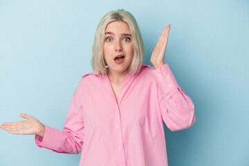 Young caucasian woman isolated on blue background holds copy space on a palm, keep hand over cheek. Amazed and delighted.