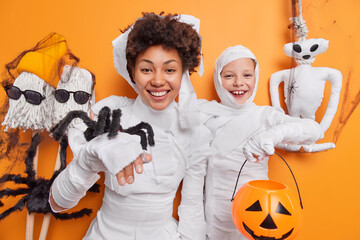 Positive optimistic Afro American woman and her little daughter hold halloween attributes smile gladfully feel carefree believe in mystery isolated over orange background. Family celebration concept
