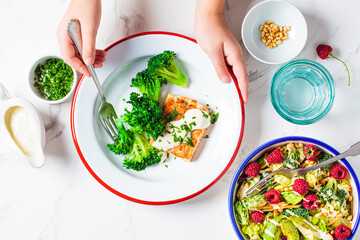 Children's hands eating fish with creamy sauce and broccoli in a white plate. Baby food concept.