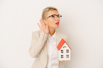 Young real estate agent woman holding a home model isolated on white background trying to listening...