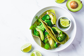 Green vegan tacos with broccoli, avocado and green flat bread in white bowl, white marble background. Vegetarian fast food.