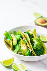 Green vegan tacos with broccoli, avocado and green flat bread in white bowl, white marble background. Vegetarian fast food.