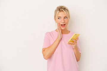 Young Russian woman holding mobile phone isolated on white background is saying a secret hot braking news and looking aside