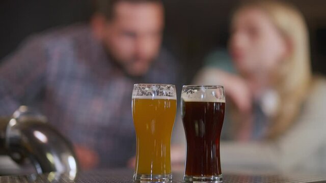 Beer glasses with pale and dark lager on bar counter and blurred Caucasian couple entering sitting down talking. Unrecognizable man and woman dating chatting in pub indoors. Lifestyle and relationship