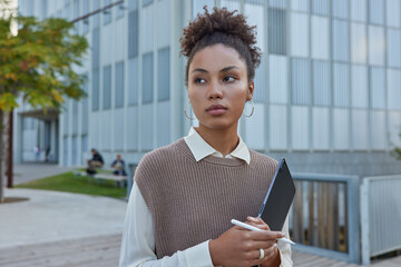 Pensive young woman with curly hair creats graphic content holds modern digital tablet and stylus...