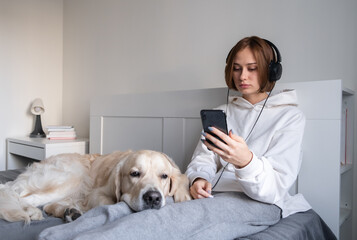 An upset girl sits with a dog on the bed, looks at the phone and listens to sad music. Teenage depression.