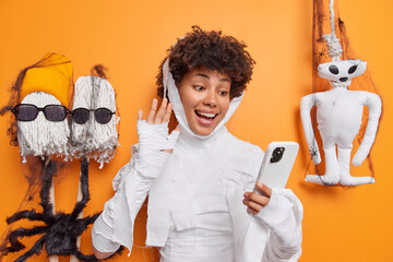 Happy young African American woman waves hello gesture in smartphone camera greets friend distantly celebrates halloween poses against orange background. Mysterious holiday on 31st of October