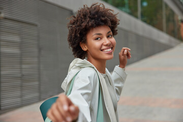 Carefree happy Afro American woman returns in good mood after fitness training carried rolled up karemat dressed in anorak looks with joyful expression poses outside against blurred background © WHstudio Leushin N