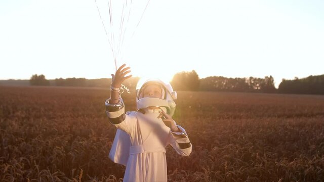 Little young happy child boy wearing astronaut uniform and helmet cosmonaut with balloons walking in sunset field nature. Space man, Kid sky big dream flying, childhood freedom, travel, future concept