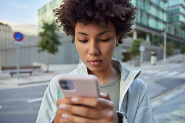 Horizontal shot of young Afro American woman with curly hair sends message on smartphone uses cellphone app strolls against urban street dressed in casual clothes focused in screen of cellular