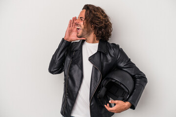 Young biker caucasian man holding a motorbike helmet isolated on gray background shouting and...